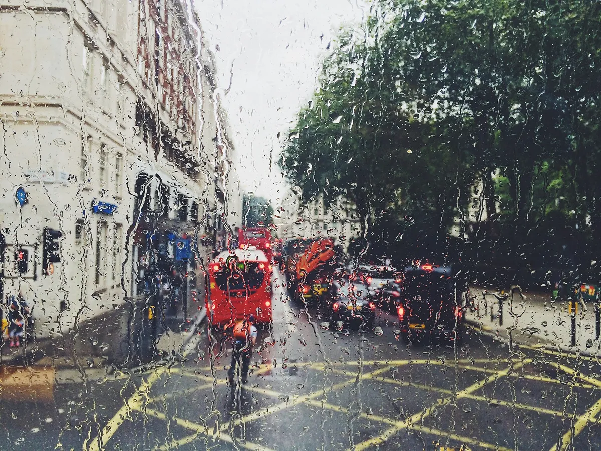 A London street in daylight seen through a rain-covered window, with a red bus and cars with their brake lights on